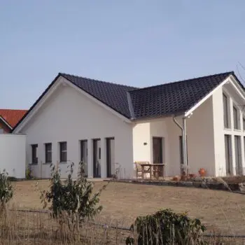 Foto eines weißen Bungalows mit dunklem Dach und mehreren Fenstern. Der Garten ist im Vordergrund.