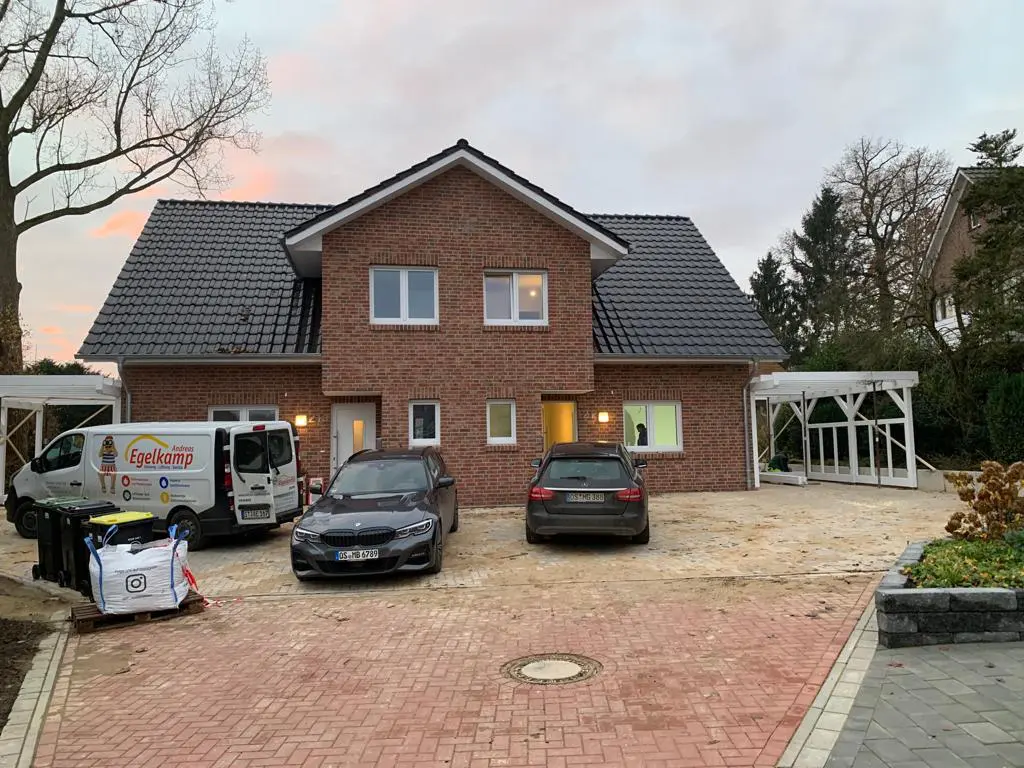 Ein zweistöckiges Backsteinhaus mit dunklem Dach, geparkten Autos und einem Lieferwagen vor dem Haus.