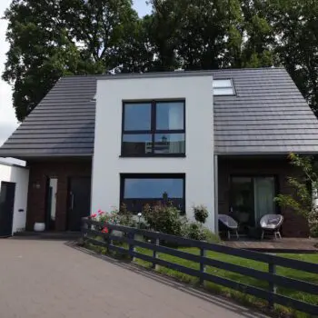 EFH-1: Modernes Einfamilienhaus mit dunklem Ziegeldach, weißen Fassadenelementen und großen Fenstern, umgeben von Grün.
