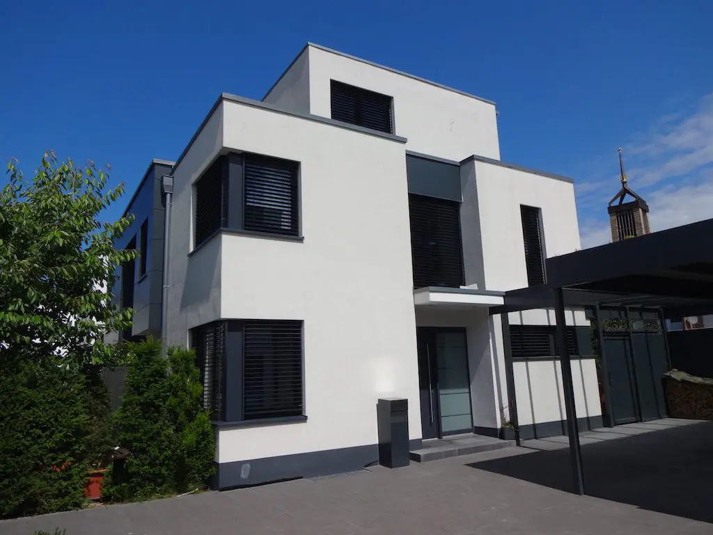 Modernes EFH mit Flachdach, weißen Fassaden und dunklen Fensterläden unter blauem Himmel.