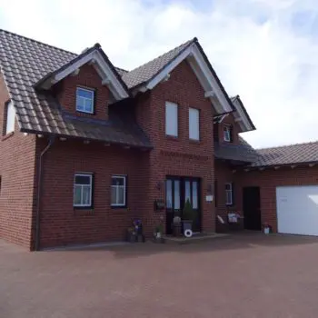 Ansicht eines EFH: Zweistöckiges Einfamilienhaus mit rotem Backstein, dunklem Ziegeldach und Garage an einem sonnigen Tag.
