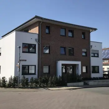 Ansicht eines modernen Mehrfamilienhauses (MFH) mit Fassade aus Backstein und weißen Elementen, umgeben von Grün.