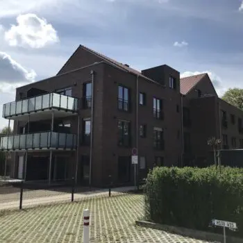 Mehrfamilienhaus mit Backsteinfassade, Glasbalkonen und gepflastertem Parkplatz im Freien.
