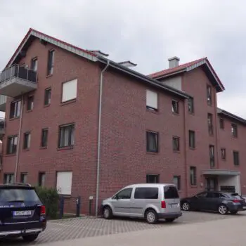 Mehrfamilienhaus (MFH) aus rotem Backstein mit Balkonen und geparkten Autos vor dem Gebäude.