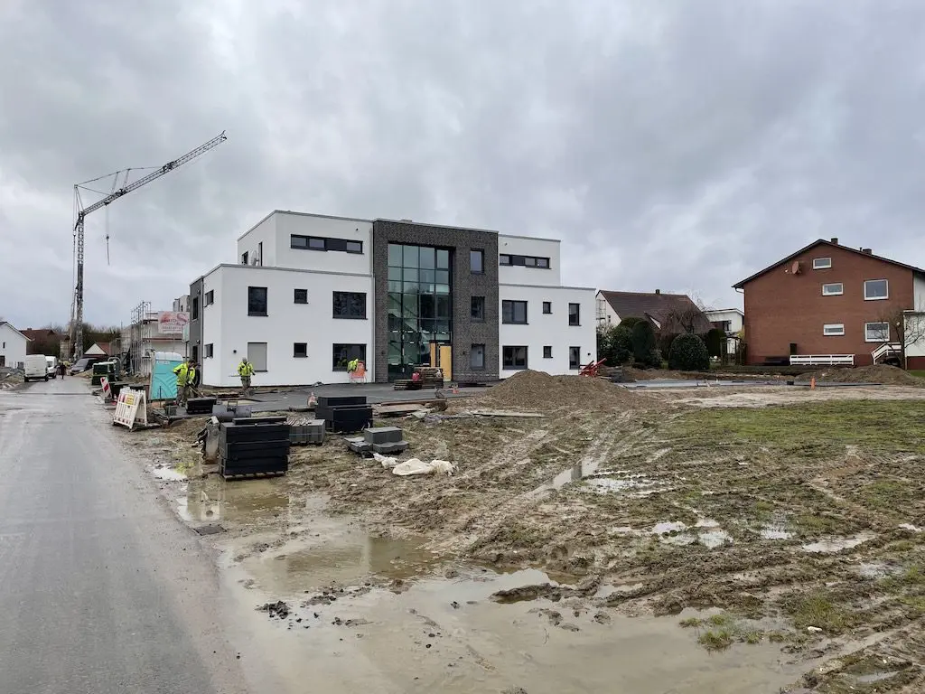 Mehrfamilienhaus im Bau: Kran, Arbeiter, Baustelle und neues Wohngebäude unter bewölktem Himmel.