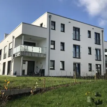 Modernes Mehrfamilienhaus (MFH) mit weißen Fassaden, Balkonen und gepflegtem Grünbereich unter blauem Himmel.