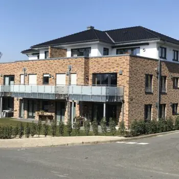Mehrfamilienhaus (MFH) mit Ziegelsteinfassade, Balkonen und dunklem Dach an einem sonnigen Tag.