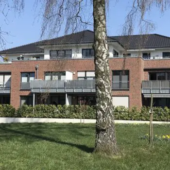 Mehrfamilienhaus mit Ziegelmauerwerk, dunklem Dach und Balkonen vor blauem Himmel.