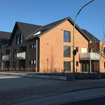 Mehrfamilienhaus mit Backsteinfassade, dunklem Dach und Balkonen an einem sonnigen Tag.