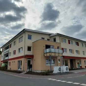 Mehrfamilienhaus mit sozialer Einrichtung, helle Fassade, Balkone und roter Zierstreifen unter bewölktem Himmel.