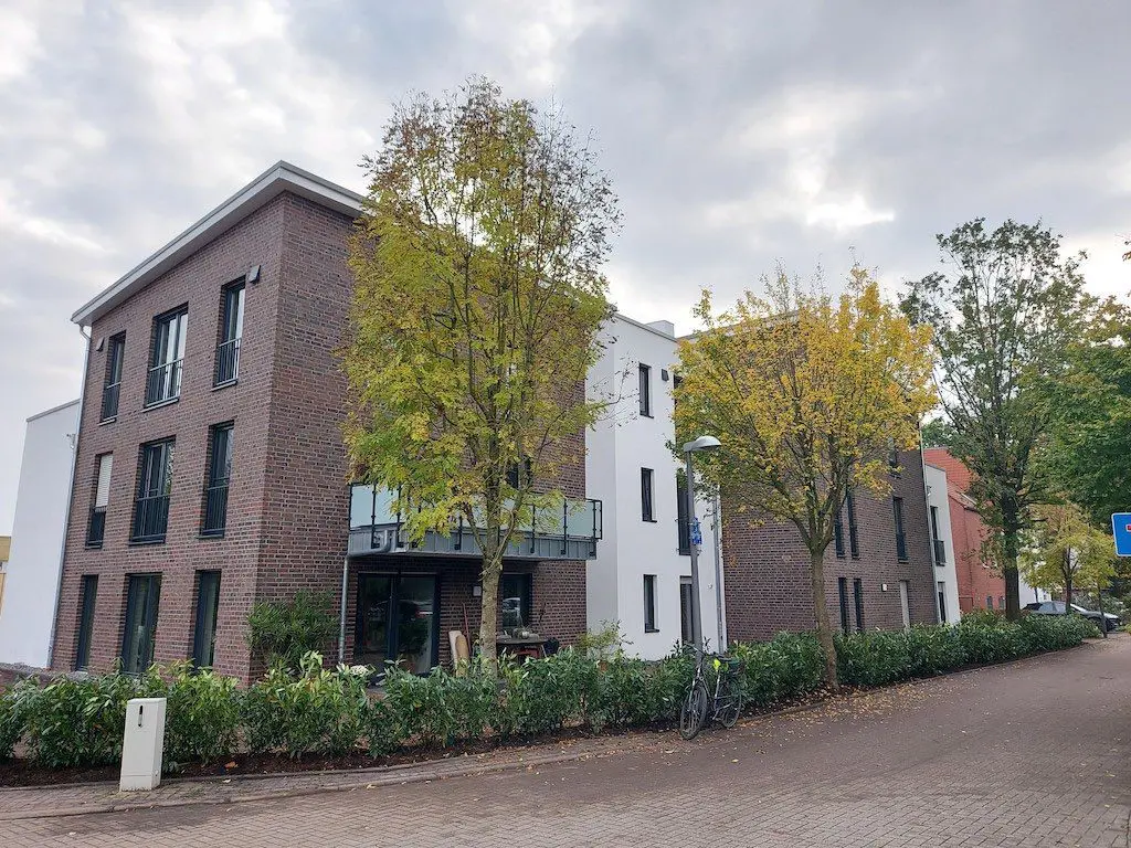 Außenansicht eines modernen Mehrfamilienhauses mit Backsteinfassade und begrüntem Vorgarten bei herbstlichem Wetter.