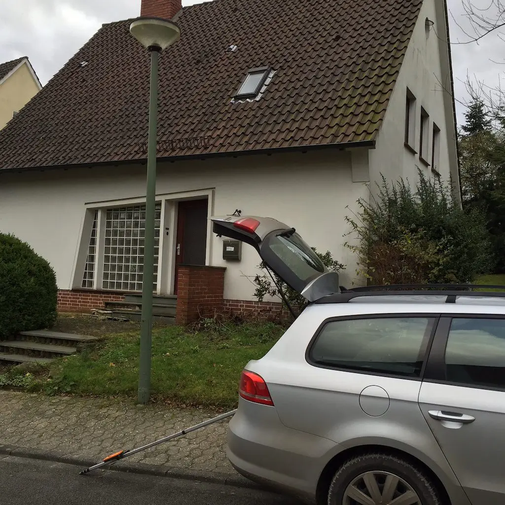 Ein Auto mit geöffneter Heckklappe vor einem Altbau mit Ziegeldach und einem Fenster mit Glasbausteinen.