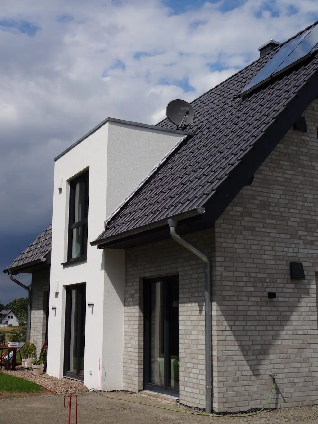Nahaufnahme eines 'Lotte' Einfamilienhauses: Graue Ziegelfassade, moderne Architektur mit weißen Putzelementen und dunklen Fensterrahmen.
