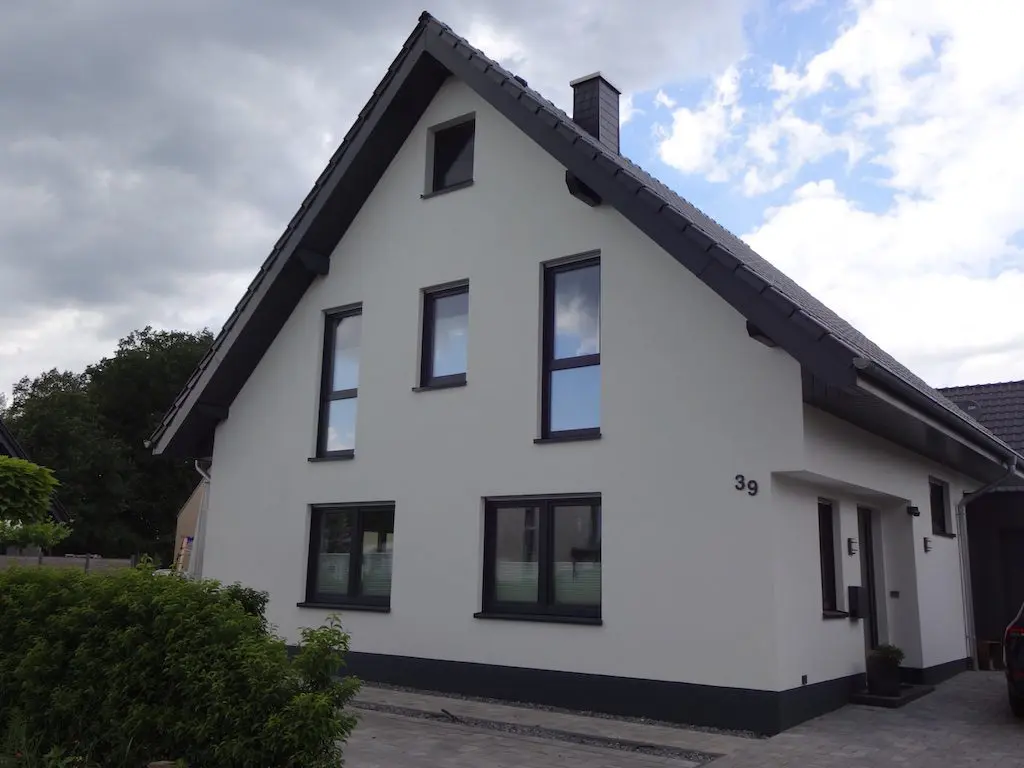 Ansicht eines Einfamilienhauses mit grauen Fensterrahmen und Satteldach unter bewölktem Himmel.