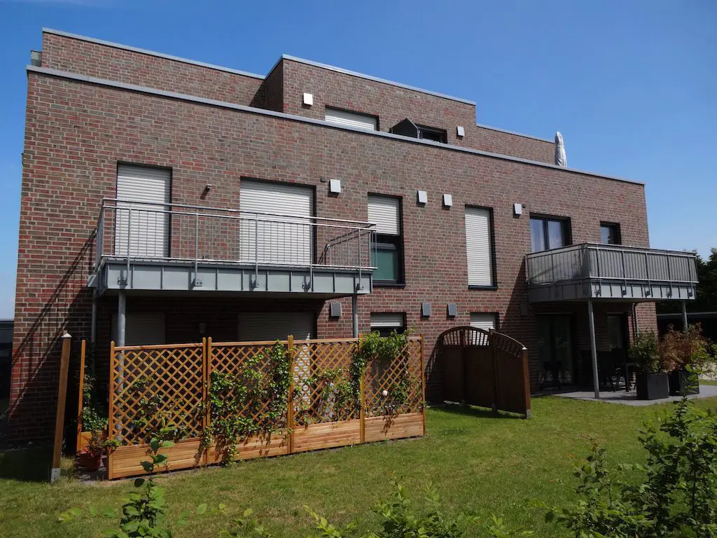 Mehrgeschossiges Gebäude mit Ziegelverblendung, Balkonen und hölzernen Sichtschutzelementen im Gartenbereich unter strahlend blauem Himmel.