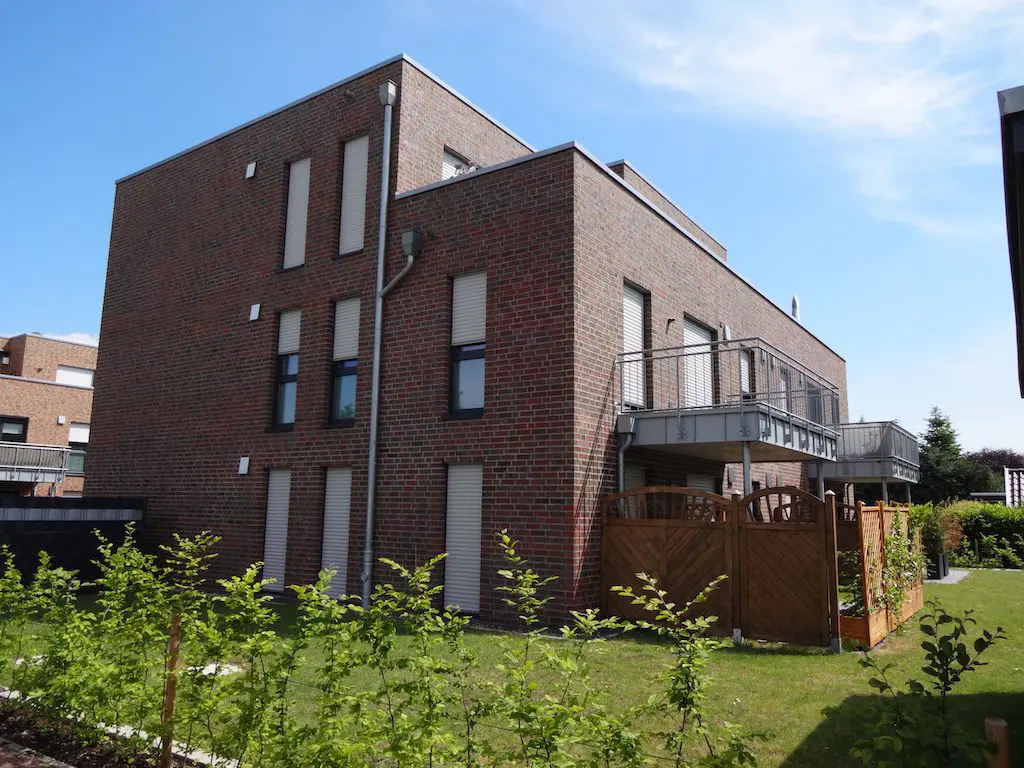 Zweifamilienhaus mit roter Backsteinfassade, zwei kleinen Balkonen und gepflegtem Garten vor blauem Himmel.