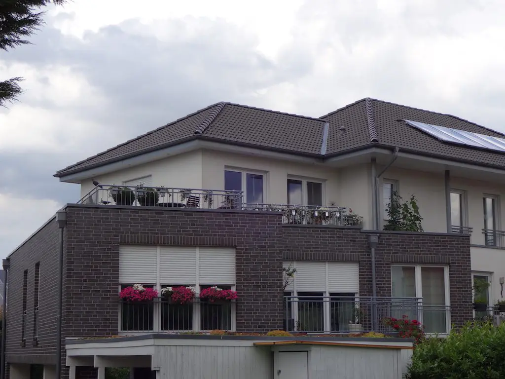 Mehrfamilienhaus mit Ziegelmauerwerk, Balkonen, Dachterrasse und Blumenkästen an den Fenstern, graues Ziegeldach.