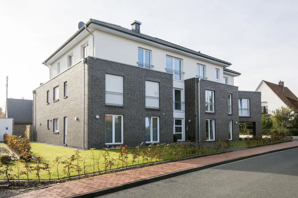 Mehrfamilienhaus mit dunklen Ziegeln und weißen Fassadenelementen, Rasenfläche im Vordergrund.