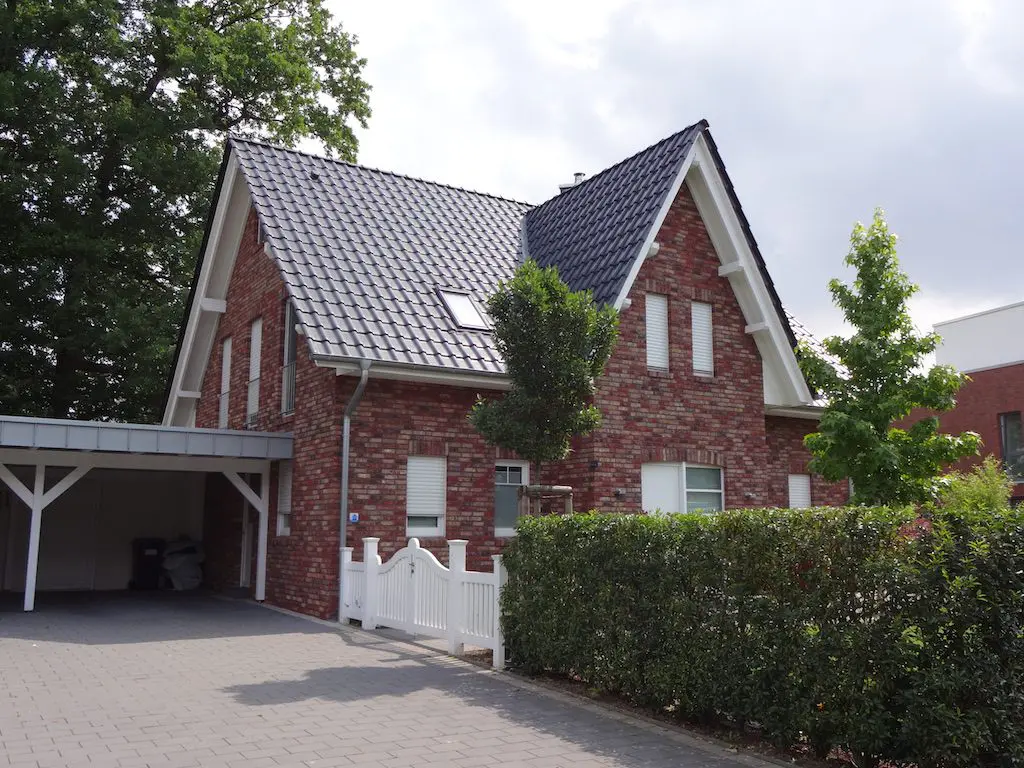 Ansicht eines Einfamilienhauses (EFH-2) mit Carport, roter Backsteinfassade und gepflegtem Vorgarten hinter weißem Zaun.