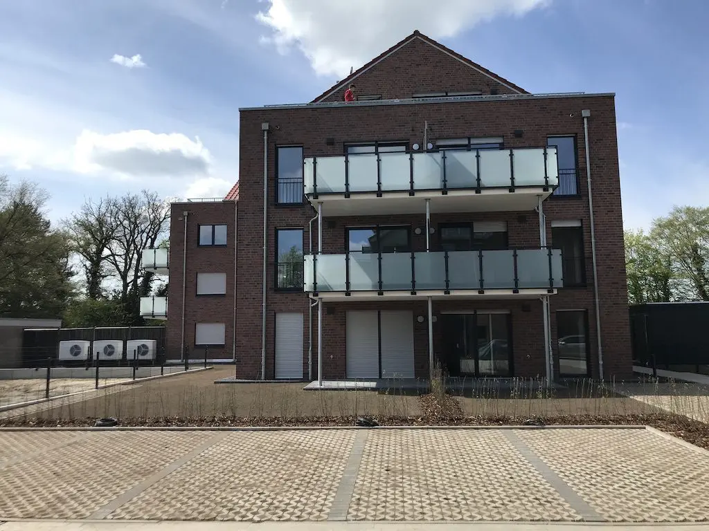 IMG_1197.jpg: Mehrfamilienhaus mit roten Ziegeln, Balkonen mit Glasgeländer und Parkplatz im Vordergrund.