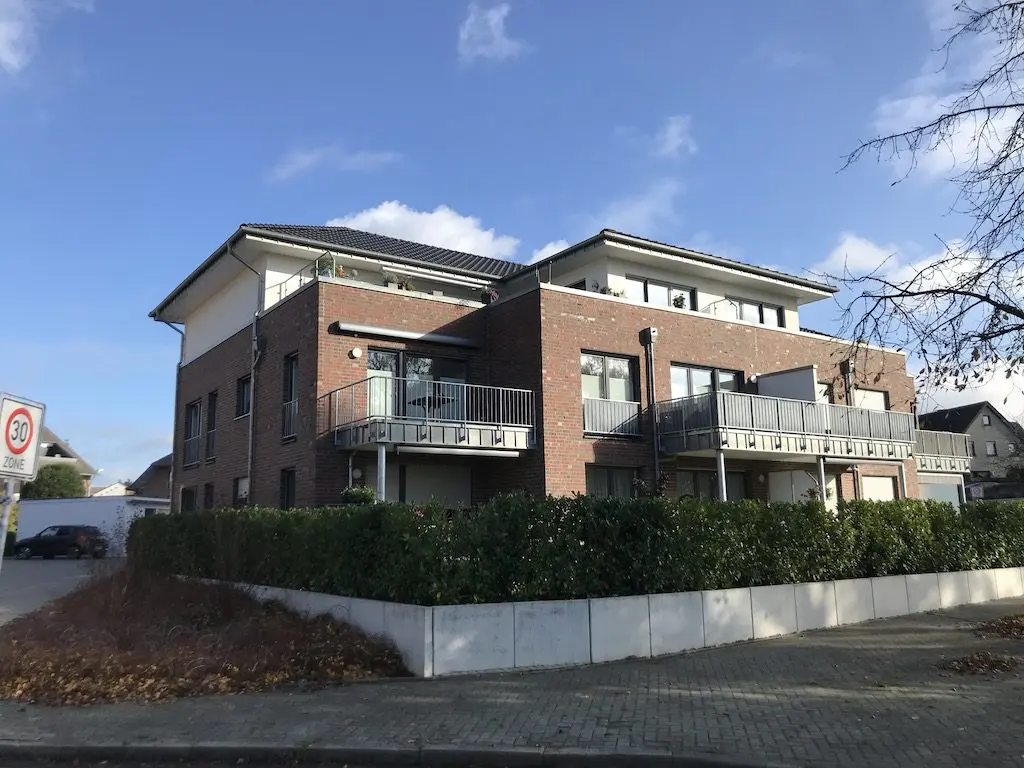 Mehrfamilienhaus mit Backsteinfassade, Balkonen und gepflegtem Grünstreifen unter blauem Himmel.
