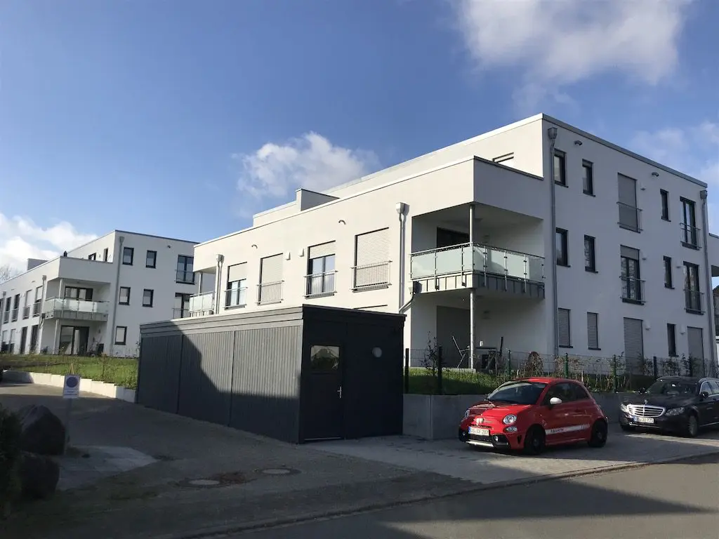 Moderne weiße Wohnhäuser mit Balkonen, Garage, roter und dunkler PKW unter blauem Himmel.
