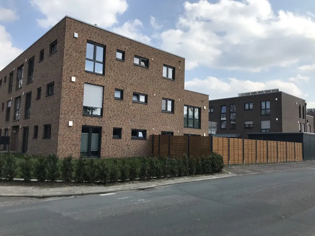 Moderne Stadthäuser mit Ziegelmauerwerk und Flachdach, umgeben von einer hölzernen Grundstücksbegrenzung, gesehen von der Straßenebene.