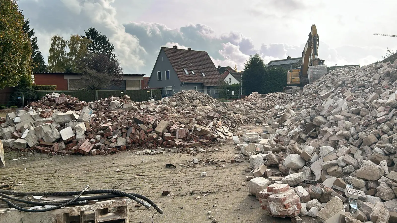 Bagger auf Baustelle vor Trümmerhaufen aus Ziegelsteinen und Beton, im Hintergrund Wohnhäuser unter bewölktem Himmel.