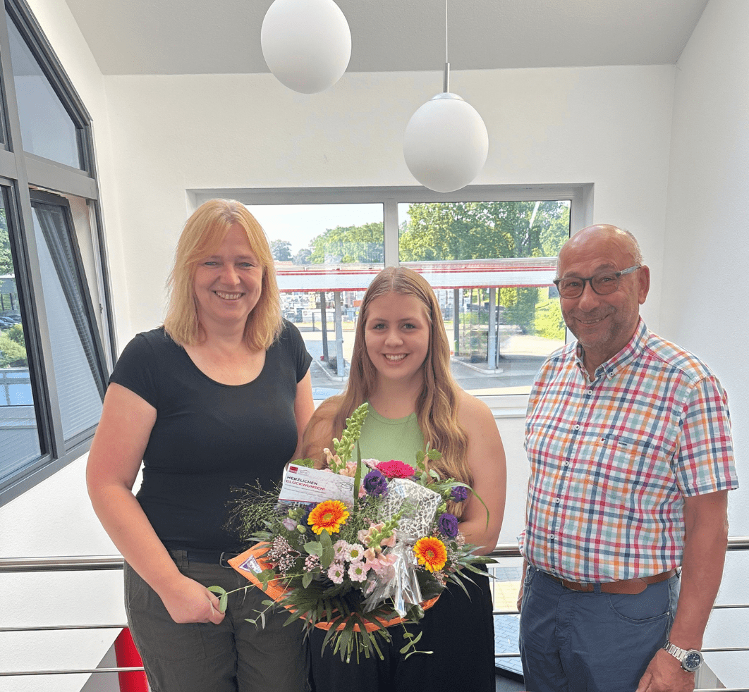 Absolventin mit Blumenstrauß und Glückwunschkarte zur bestandenen Abschlussprüfung posiert lächelnd mit zwei Kollegen